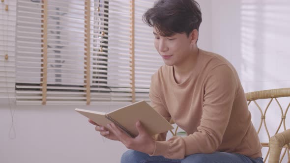 At home, a handsome young Asian businessman in casual clothing reads a book on the couch.