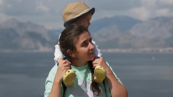 Mother Puts Little Son on Shoulders Standing Against Sea