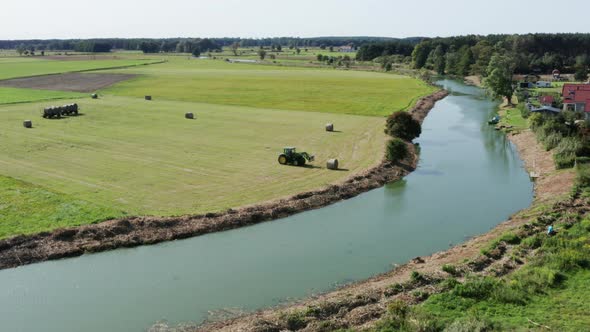 Aerial agricultural shot of a tractor collecting bales of hay by the curvy river in a farm village.