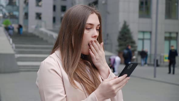 Shocked Young Woman Looking at Smartphone Standing Outside Office Building