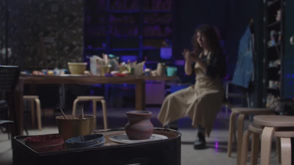 A Fresh New Pot Spinning on the Wheel and a Young Woman Sitting By the Table with Clay in Her Hands