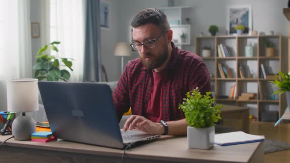 Young Male Freelancer in Red Shirt Sitting at Table and Working on Wireless Laptop