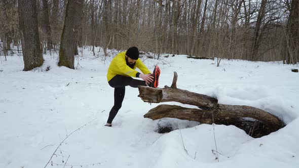 Fit Bearded Sports Man in Yellow Coat Stretching Legs before Running in Winter Forest at Snowfall