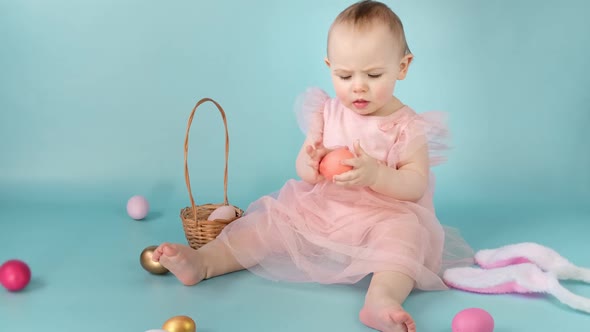 Happy Baby Girl in Bunny Ears Headband Sitting on Blue Studio Background and Playing Easter Eggs