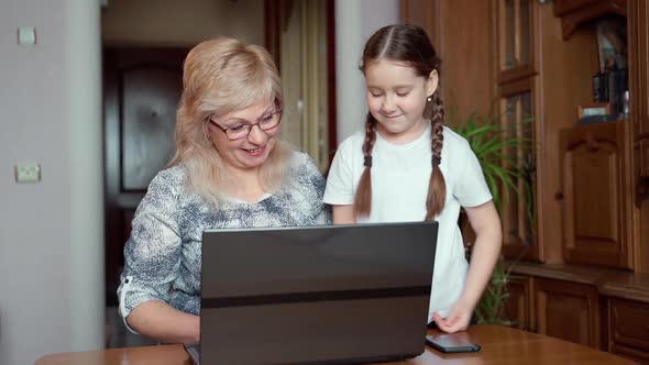 Little young school age girl teaching grandmother using laptop notebook tech at home. Slow motion