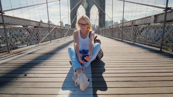 Young Woman Sits on the Brooklyn Bridge in New York, Uses a Smartphone