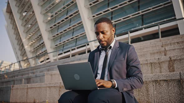 Concentrated African American Businessman in Suit Typing Project on Laptop Sitting on Stairs