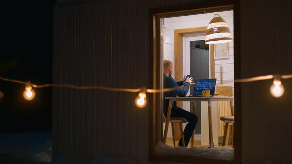 View From the Outside Through the Window a Man Who Works on a Laptop at Home Takes a Smartphone and