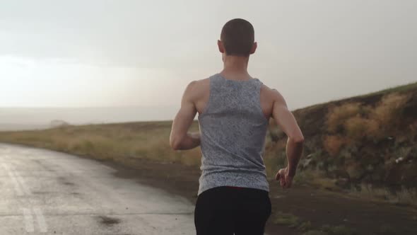 Man of Athletic Build Runs Along Road in Countryside During the Rain Flash of Lightning View Back