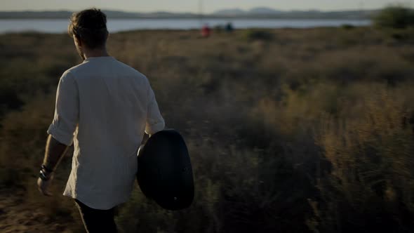 Spanish Hipster Man with a Guitar Walking Through a Grassy Field Towards Sunset Following Gimbal