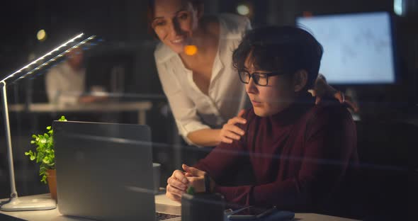 Female Executive Helping Young Stressed Employee Sitting at Desk Working Late in Office