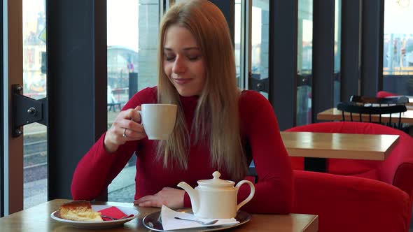 A Woman Sits in a Cafe with Breakfast on the Table, Looks Out a Window and Drinks Tea with a Smile