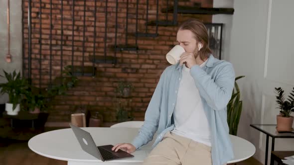 An Attractive Young Employee Having a Break Scrolling His Lapyop and Drinking Coffee