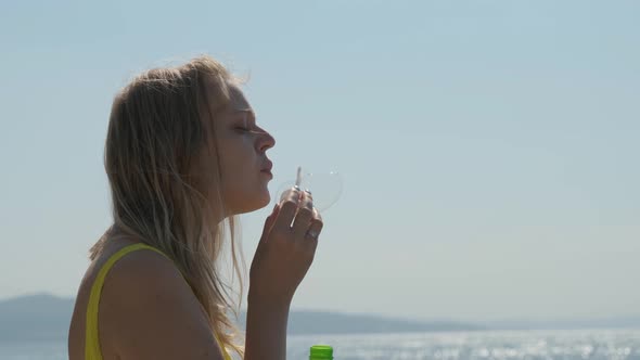 Blond Young Woman Blows Bubbles on the Background of the Blue Sea. She Enjoys This and Is Happy.