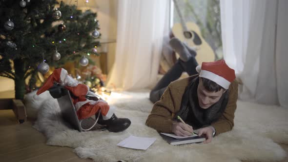 Wide Shot Portrait of Joyful Young Caucasian Man Writing Letter To Santa at Home