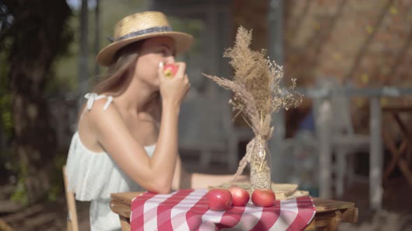 Young Rural Girl in a Straw Hat and White Dress Sitting at the Small Table with Vase with Decorative