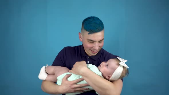 A Young Father with Blue Hair Holds a Baby in His Arms. Alternative Person Cradles a Child on a Blue