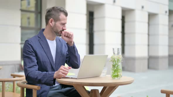 Man Coughing While Using Laptop Sitting in Outdoor Cafe