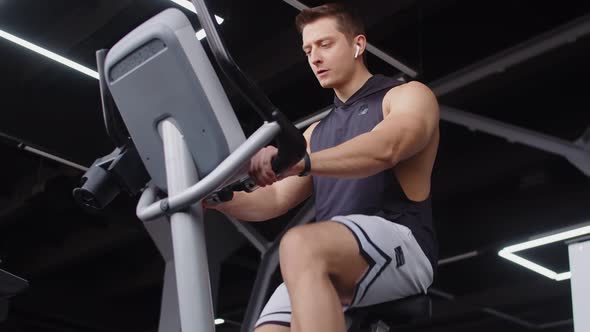 Attractive Man with Headphones of Athletic Build Trains Pedaling on an Exercise Bike in Gym Front