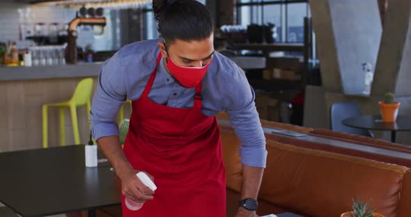 Mixed race male cafe worker wearing face mask disinfecting table