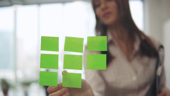 A Young Woman in a Business Suit Sticks Sticky Sheets with Reminders on a Glass Partition