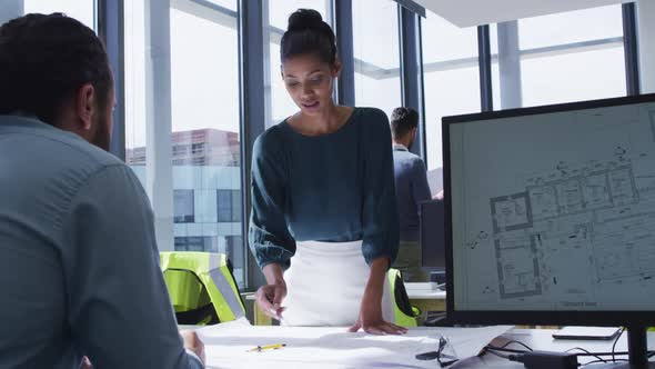 Two diverse male and female colleagues looking at architectural blueprints on desk and talking