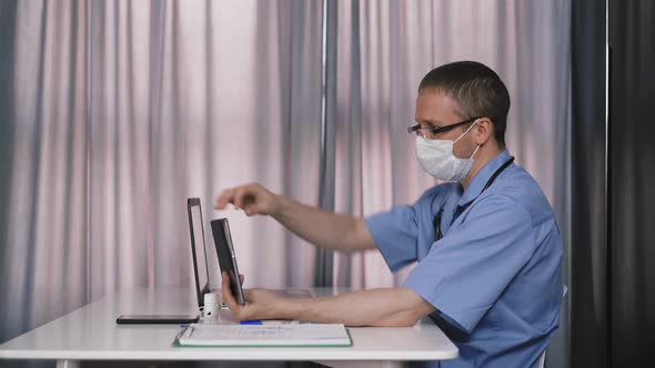 Doctor in Mask Has Conference Via Tablet and Laptop at Table