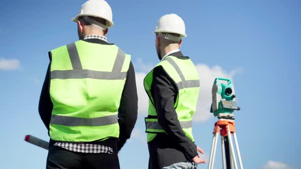 Two Caucasian Young Men in Uniform Standing at Theodolite Outdoors Talking Pointing Looking Away