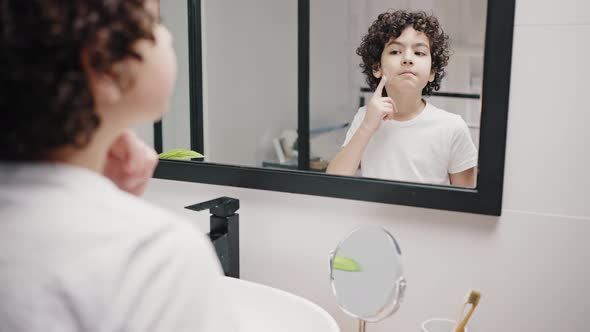 Cute Boy with Curly Hair Puts Cream on Cheeks in Bathroom