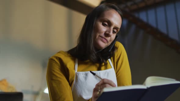 Caucasian female jeweller in workshop wearing apron, sitting at desk, having call and taking order