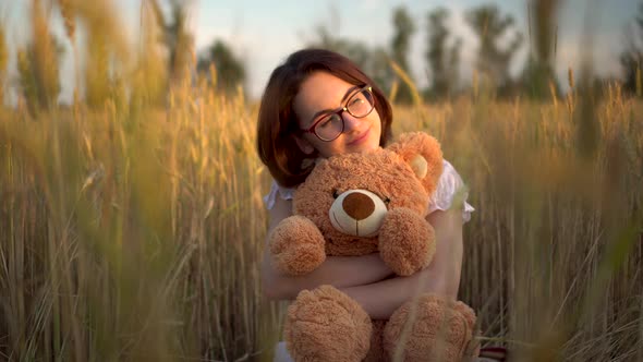 A Young Woman Sits in a Dried Wheat Field with a Teddy Bear. Girl Hugs a Teddy Bear in Hands Front