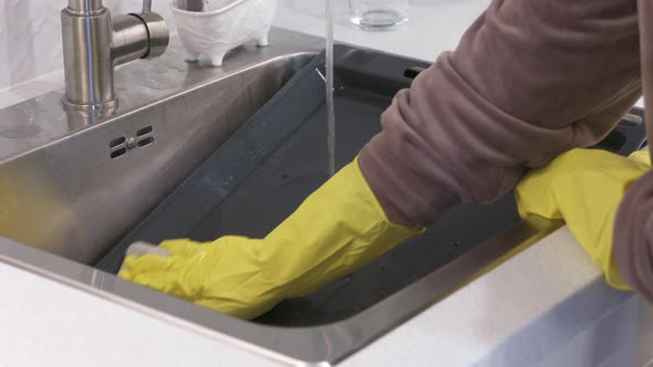 Woman Wearing Rubber Gloves Cleaning Oven Baking Sheet in Kitchen Sink Under Running Water