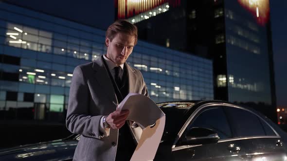 Handheld Portrait of a Young Businessman Standing Near a Black Car and Reads Financial Documents Top