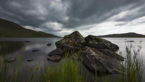 Motion time lapse of lake with grass and large rocks in the foreground on a dark cloudy summer day i