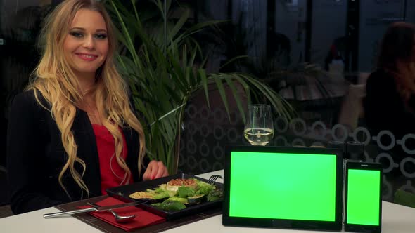 A Woman Sits at Table in Restaurant, Smiles at the Camera - a Tablet and a Smartphone - Green Screen