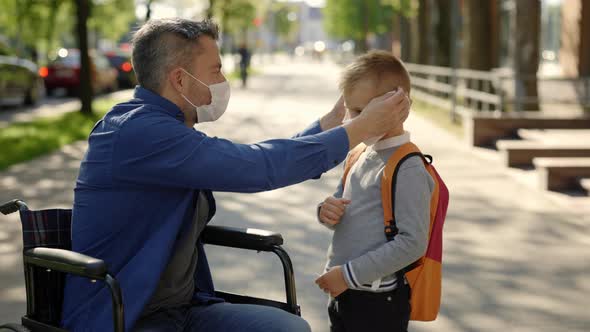 Disabled Father in Mask Sitting on the Wheel Chair Wearing Mask to His Son Before School