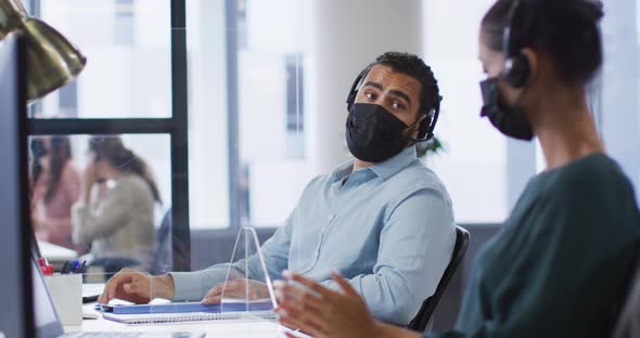 Diverse male and female colleague in face masks and headsets talking at desk with sneeze shield