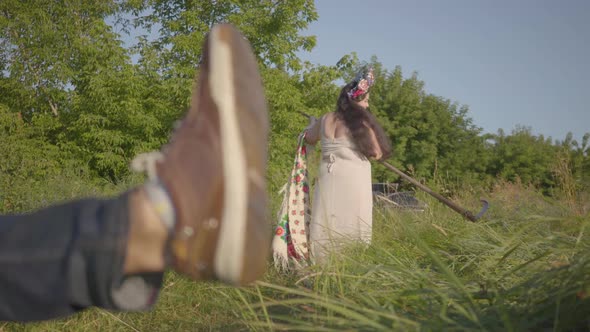 Foot of a Young Man Resting on the Ground in the Foreground. Plus Size Woman Finishing To Mow the