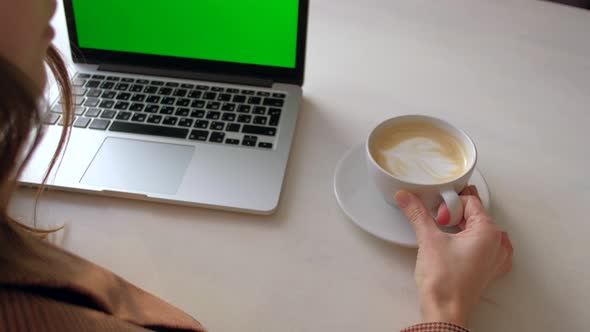 Back view - woman sitting at table drinking coffee and looking at laptop.