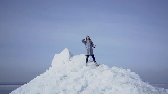 Blond Woman with Long Hair in Warm Coat Standing on the Top of Glacier Throwing Up Her Hands