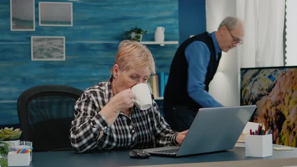 Older Mature Business Woman Using Laptop Sitting at Workplace Drinking Coffee