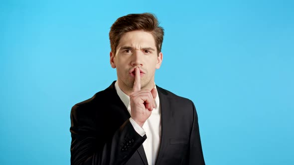 Serious Business Man in Professional Suit Holding Finger on Lips Over Blue Background. Gesture of