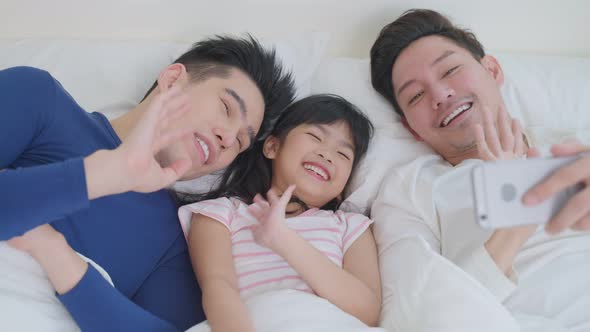 Asian LGBT gay family use mobile phone with daughter video call on bed.