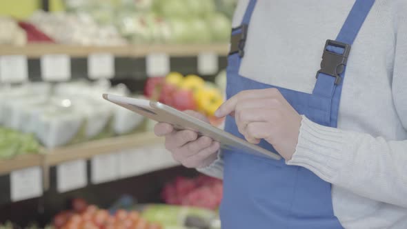 Unrecognizable Male Grocery Worker in Uniform Walking Along Shelves and Using Tablet. Adult Man