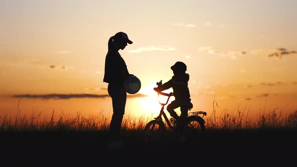 Silhouette of Mother Teaching Little Son to Ride a Bike at Meadow During Sunset