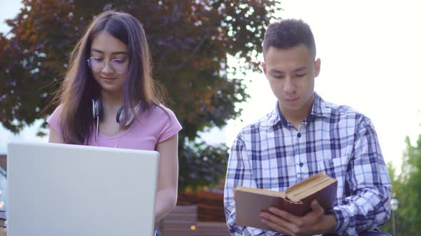 Young Male Asian Student with Book and Young Asian Woman with Laptop in Park