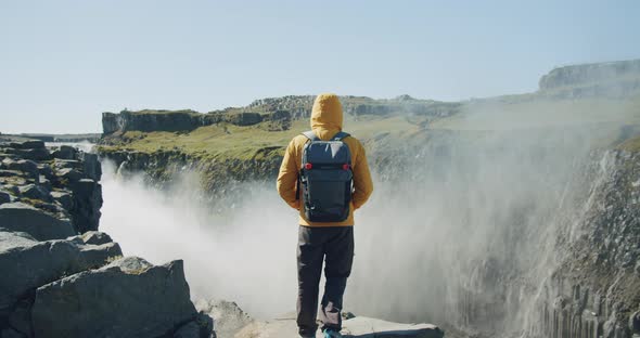 Rear View of Man in Yellow Jacket and Backpack Standing at Cliff Edge Looking at Detifoss Waterfall