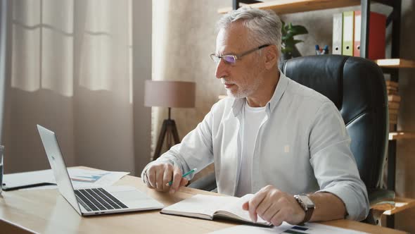 Mature Male is Typing on Laptop Computer and Writing in Notebook While Sitting at Table Working at