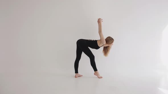 A Slender Young Girl in a Black Top and Leggings is Engaged in Stretching in a White Studio and