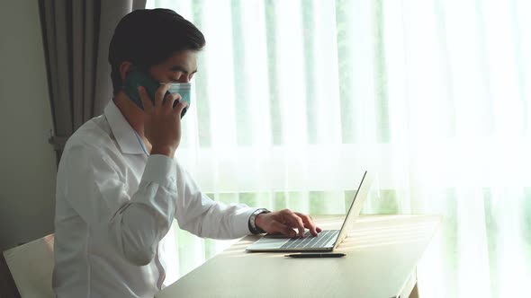 Asian man in mask taking a call on his mobile phone checking information received on his laptop 